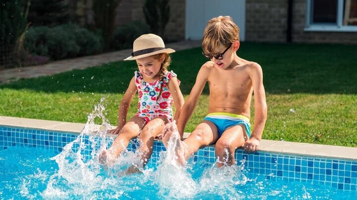 Niños chapotean sentados en el borde de una piscina comunitaria con un jardín de césped al fondo