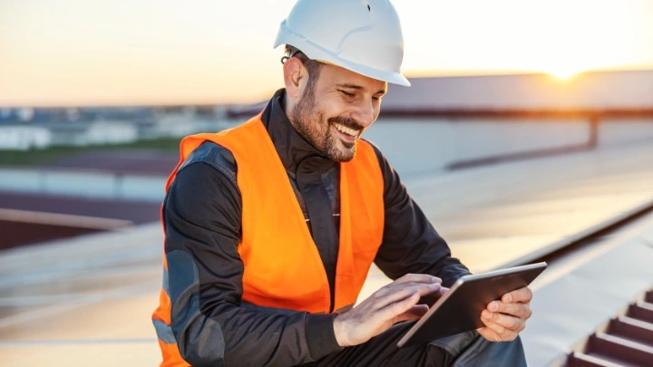 Un obrero de la construcicón sobre un tejado trabaja en una tablet frente a una bonita puesta de sol.