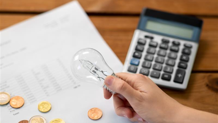 Una mano rodeada de facturas eléctricas con una calculadora. Se ven sobre la mesa bombillas y dinero.Mano con calculadora-de-facturas-de-bombillas-y-dinero.