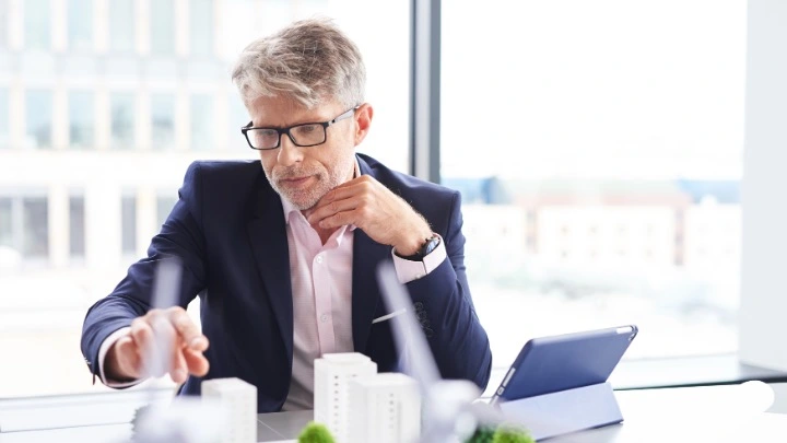 Ejecutivo de mediana edad con gafas analiza una maqueta de edificios sobre una mesa en una oficina moderna mientras utiliza una tableta.