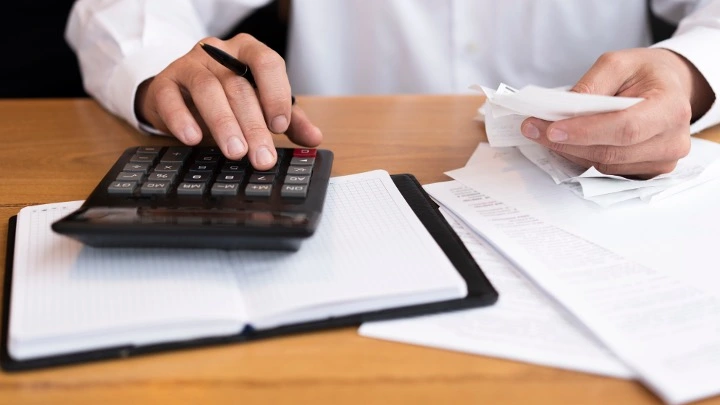 Persona utilizando una calculadora y revisando facturas o documentos financieros sobre una mesa, en un contexto de gestión económica o contable.
