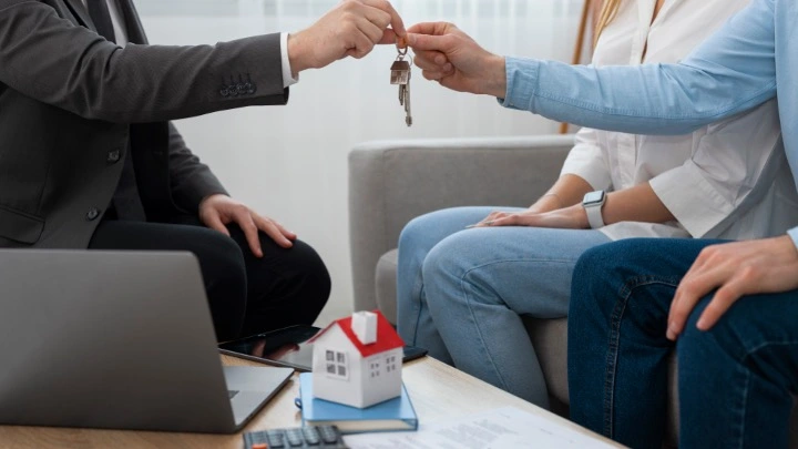 Persona entregando unas llaves a otra en un entorno profesional, con documentos y un modelo de vivienda sobre la mesa, simbolizando una operación de alquiler o compraventa inmobiliaria.