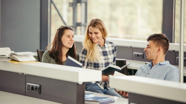 Tres estudiantes sentados en un aula moderna, conversando y estudiando juntos mientras revisan libros y apuntes sobre una mesa.