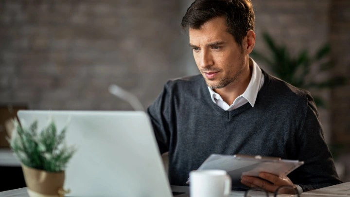 Hombre revisando documentos mientras trabaja con un ordenador portátil, en un entorno de oficina relacionado con la gestión y el control financiero.